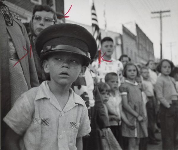 A boy wearing a peaked cap is looking to his right. Behind him are rows of standing people, and an American flag. Caption reads: "As the marchers flashed by, Ronald Moss, 7, of Kaukauna watched in awe."