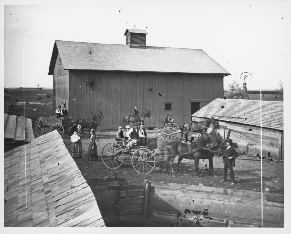 Barnyard, with what appears to be a pigyard littered with corncobs. It is enclosed with a solid board fence and has slop troughs made from hollowed logs. Also in this farmyard view is a family in carriages and on horseback. A mower, dump rake, windmill and barn are in the background.