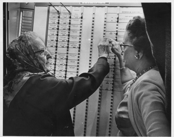 An older woman and a younger woman talking in front of a voting machine. Caption reads: "HASN'T MISSED AN ELECTION — Mrs. Mary Sturm, 71, of 820 E. Ogden av., was briefed on the use of a voting machine by an election supervisor, Mrs. Harry Sanders, 1544 N. Humboldt av., Tuesday at the Cass Street school. Mrs. Sturm said she had not missed voting in an election since women were given the right to vote."