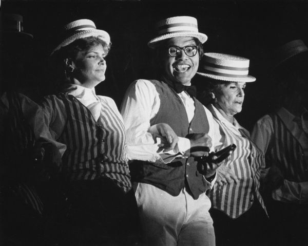 A man has linked arms with two women and is posing looking at the camera. The group is wearing costumes consisting of vests and straw boater hats. Caption reads: "<b>It's time for Follies!</b> Three members of the cast for 'Night Club Nostalgia,' the annual fund-raising follies for the Waukesha Symphony Orchestra, performed Thursday night, with Sam Arreola singing and dancing to 'Alexander's Ratime [sic] Band.' The follies continue tonight and Saturday night at Waukesha North High School."