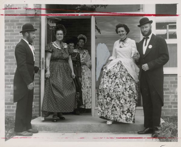 Two men and four women posing outdoors in costume in the open doorway at the front of a building. Caption reads: "<b>STEPPING DOWN</b> from the entry of the new St. Croix county highway department building, scene of Tuesday's centennial activities, Hammond folks in costume furnish a contrast between the past and present. From left they are Clyde Rasmussen, Mrs. John Oslun, Mrs. Henry Schieven, Mrs. Ted Riley and Ted Riley."