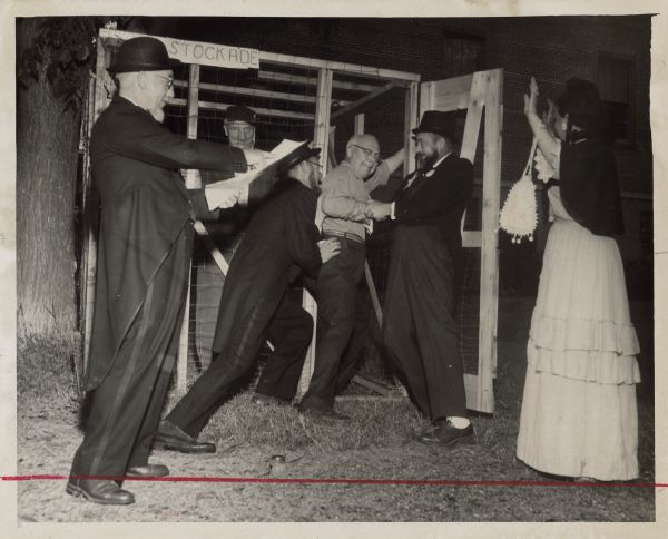 View of an outdoor performance with a tree on the left, and a brick building in the background. Two men in suits are attempting to push a third man into a stockade, in which another man is standing. A fifth man is standing on the left holding a document and pointing at it, and a woman on the right is holding up her hands. Caption reads: "<b>A BEARDLESS RESIDENT</b> gets thrown into the 'stockade' with another culprit. Reading the citation is John Oslun. Fred Hartwig looks out of the cage while Henry Schieven and Donald Grinnel shove protesting Edwin Larson through the door. Mrs. Donald Grinnell, at right, expresses horror at the event."