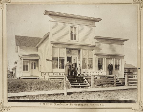 Exterior view of the store with a false front. A sign along the porch on the left reads: "Millinery" and a sign in the center reads: "Wm. Dittberner, Merchant Tailor". Standing in front of the building from left to right are: Mrs. William Dittberner, Mary Dittberner, Lena Dittberner and William Dittberner. The woman and two girls are standing together with a small dog at their feet in front of the entrance to the Millinery shop, and William Dittberner is standing on the right in front of another door for the Merchant Tailor shop. A sign attached on the left side corner of the shop reads: "'Jennie June' Sewing Machine". Hats are displayed inside the front windows, and posters with hat designs cover some of the windows around the main doorway. A window to the right of William Dittberner has a poster of men's suit designs. Another entrance to the building is set back on the left with a small roofed entryway. Buildings are in the background on the left and right.