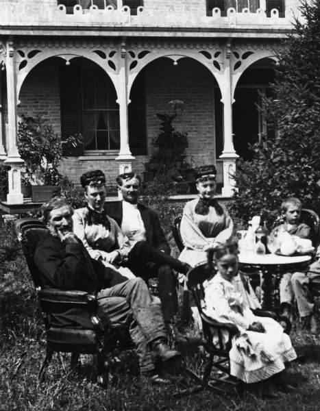 Detail from original of part of the family. Original caption reads: "Family seated around table in yard with baby on grass and brick house with porch trim and shutters in background. This house is said to resemble the J. Hill house on Hubble Street in Black Earth, Wisconsin."