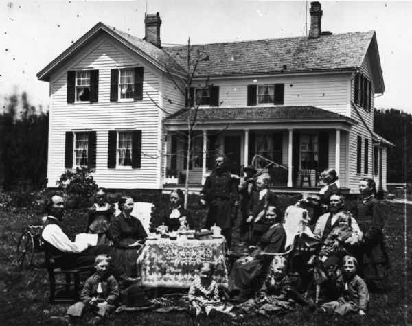 Detail from original of the family. Original caption reads: "Norwegian family posing with their household possessions, in front of their home. The man dressed in military uniform is a Civil War veteran. Madison, Wisconsin (area), ca. 1870-1879. The family is in a yard around a table adorned with a lace cloth. Coffee cups, figurines and books are on the table. On the left is a man holding a newspaper that says 'Norden.' A baby sits in a buggy behind him. In the rear is a man dressed in a military uniform and, behind him, a man with a rake. On the right is a woman with a tray of beverages and, in front, a rocking horse."