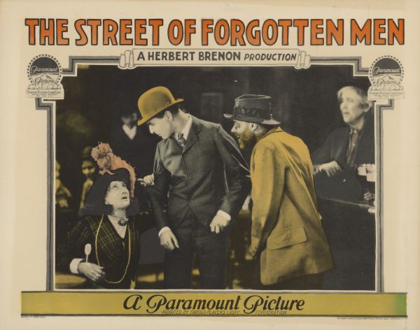 Lobby card for the 1925 film "The Street of Forgotten Men". An older woman, sitting and wearing a nice dress and hat, is grabbing the arm of a young man standing next to her. Another man, wearing shabby clothes and a beard, is smiling and looking at them.