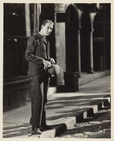 Easy-Money Charley, played by Percy Marmont, stands on the street curb in front of a light pole in the 1925 film "The Street of Forgotten Men". He holds his hat in his hands.