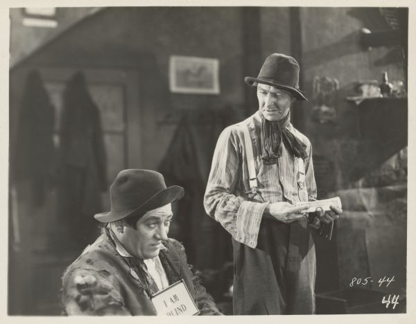 Easy-Money Charley (Percy Marmont) looking down at a seated Bridgeport White-Eye (John Harrington) in a scene from the 1925 film "The Street of Forgotten Men". The men are both dressed in shabby clothes and White-Eye wears a "I AM BLIND" sign around his neck. Charley is holding a folded dollar bill.