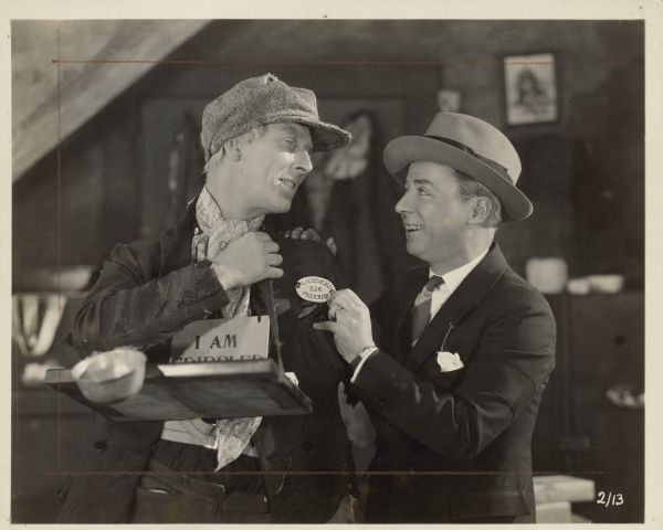 Film director Herbert Brenon holds a "LICENCED PEDDLER" pin to Percy Marmont's lapel. Marmont is dressed as a crippled peddler for the 1925 film "The Street of Forgotten Men".