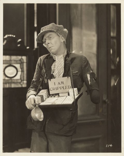 Percy Marmont, as Easy-Money Charlie in the 1925 film "The Street of Forgotten Men", standing on the street with a tray of items and a sign reading: "I AM CRIPPLED" hanging from his neck. He is holding a small cup in one hand; his other hand is "missing".