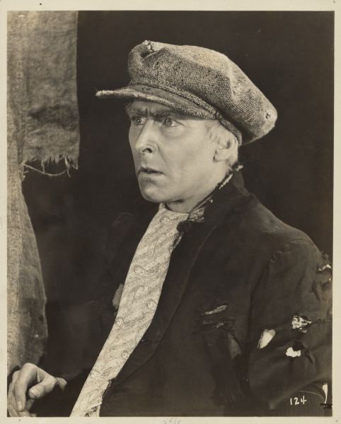 Easy-Money Charley, played by Percy Marmont, has a look of consternation on his face in a scene from the 1925 film "The Street of Forgotten Men". He wears a page boy hat and a suit jacket with many holes.