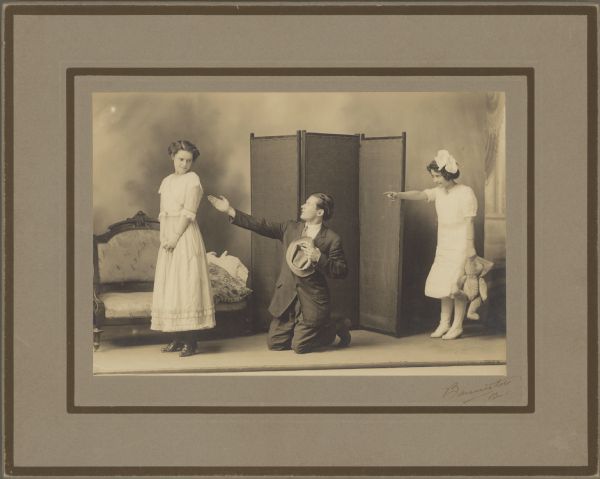 Posed scene from a play. A man kneels and reaches out to a woman, who is turned away but looking back at him. Another actress is dressed as a young child, holding a stuffed toy and pointing at the man. Caption reads: "Frank B. Wood. Senior Class Play, Neillsville H. S. — 1912".