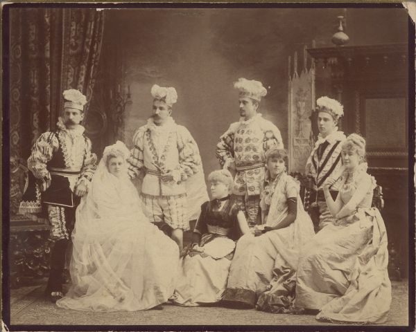 Group portrait of four men and four women, all in costume. One woman is dressed in a bridal gown. Caption reads: "'The Mistletoe Bough' 1885. Mary R. Brigham as the bride".