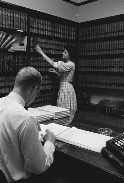 Shirley Abrahamson is standing in a library reaching for a book. A man is sitting in the foreground. 