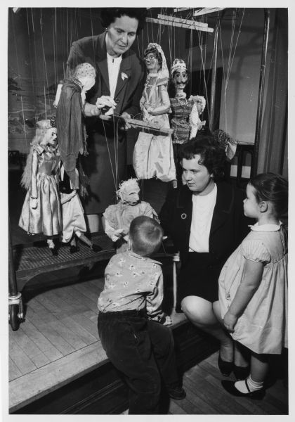 A woman standing among several marionettes, demonstrating the strings and control bars to another woman and two children. Caption reads: "<b>Two youngsters</b> learned what makes a marionette move when they visited the workshop of the YWCA Marionette Theater group at the downtown activities building, 610 N. Jackson st. Mrs. Ernst von Briesen, 1019 E. Sylvan av., Whitefish Bay, demonstrated for John Laborde, 3, of 4055 N. Stowell av., and Barbara Graham, 3, of 4050 N. Prospect av., Shorewood. Mrs. von Briesen and Barbara's mother, Mrs. Leo Graham, are members of the marionette theater group which will present 'The Shoemaker and the Elves' during an open house on Sunday." 