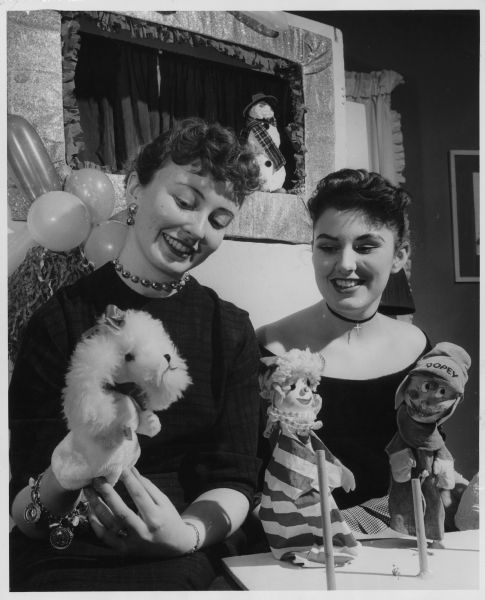 Two women smiling and looking at a hand puppet of a dog. Other hand puppets are propped up on a table in front of them, and a stage is behind them. Caption reads: "<b>Operators of a small</b> but successful puppet show business are Miss Cynthia Shove (left), 4317 N. Marlborough dr., Shorewood, and Miss Lee Hauck, 7710 N. Bell rd., Fox Point. The cousins are shown with just a few of their puppets. Behind them is the portable stage which they carry all over Milwaukee and the suburbs to perform for children's groups."