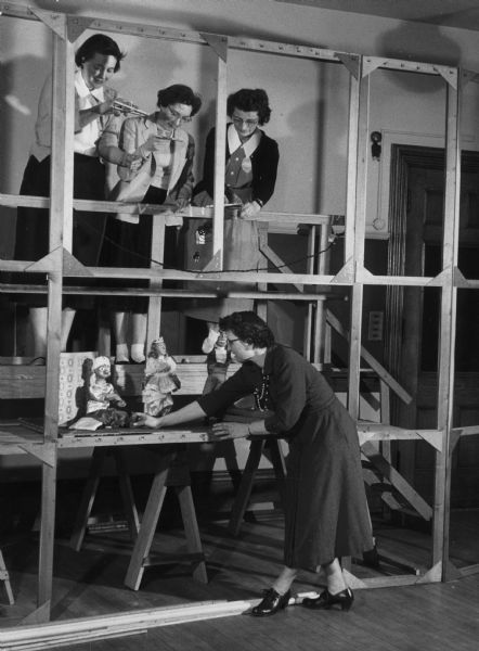 A woman adjusts a marionette on a table. Above and behind the table, three women operate horizontal ("American") control bars. Caption reads: "Mrs. Harry Hartman, assistant director of the occupational therapy department at Milwaukee-Downer college, made several adjustments on the marionettes during rehearsals. Class members tested their creations on the skelton [sic] stage. From left are Mrs. Harold Baxter, N. Cramer st.; Barbara Smith, Little Rock, Ark., and Sister Rose Kroeger, W. Kilbourn av., a Lutheran deaconess. The play was given Wednesday to prove the skill of the class."