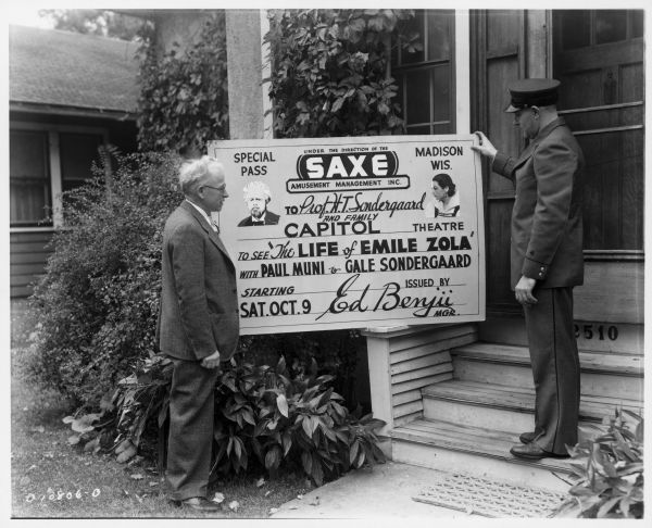 Two men ---- one in a mailman's uniform ----- standing on either side of an oversized special pass to a screening of the film "The Life of Emile Zola" starring Paul Muni and Gale Sondergaard. The pass is made out to Prof. H.T. Sondergaard and Family. Caption reads: "Madison, Wis., 1937. Mailman Merle Patton and Prof. H.T. Sondergaard holding complimentary ticket to movie starring Gale Sondergaard."