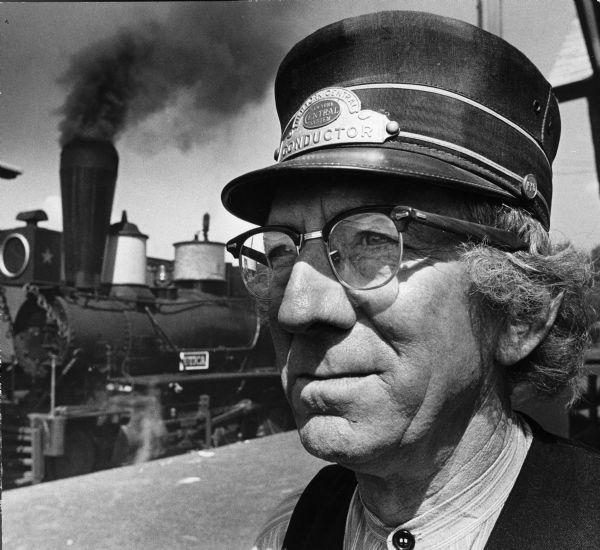 Close-up of a man wearing a conductor's hat. A train is in the background. Caption reads: "Norman Wiese, North Freedom, Mid-Century RR Museum. Was also in movie - The Immigrants." [Actual title, "The Emigrants"]