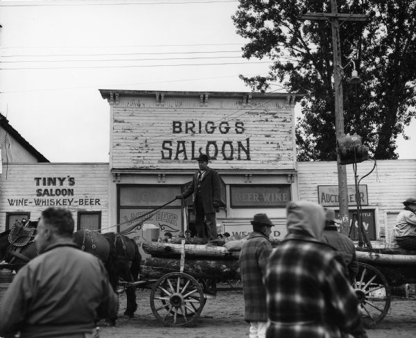 A man standing on a log cart, holding the reins for a horse team. Other people are in the foreground and background. Shopfronts for Tiny's Saloon and Briggs Saloon are in the background. Caption reads: "Saxon Wis 1961. Altered town street being used as background for production of film 'Young Man' [actual title, "Hemingway's Adventures of a Young Man"] based on stories by Ernest Hemingway. 20th Century Fox."