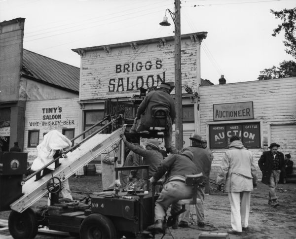 A film production crew operating a camera crane in front of a street scene, with signs for Tiny's Saloon, Briggs Saloon, and Wm. "Pop" Wiener, Auctioneer. Caption reads: "Saxon Wis 1961. 20th Century Fox film crew at work on 'Young Man' [actual title, "Hemingway's Adventures of a Young Man"] based on stories by Ernest Hemingway."  