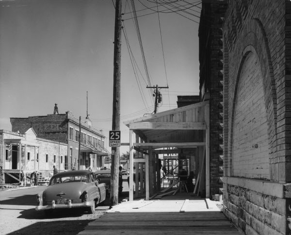 View of a town street. Wooden shopfronts are being built in front of brick buildings. A car with Michigan license plates is parked next to a telephone pole, which has a 25 mph speed limit sign. Caption reads: "Saxon Wis 1961. Town being altered to appearance of 1916 for use as location for 20th Cent rt [sic] Fox film 'Young Man' [actual title, "Hemingway's Adventures of a Young Man"] based on stories by Ernest Hemingway."
