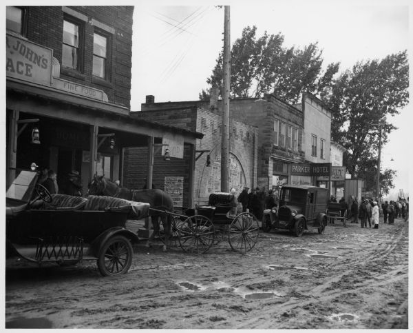 View of a street, with shop fronts, a horse-drawn carriage, and automobiles. Several people are in the background. Caption reads: "Saxon Wis 1961. 20th Century Fox Movie company reconstructed much of the town to use as a setting for a motion picture 'Young Man' [actual title, "Hemingway's Adventures of a Young Man"] based on Ernest Hemingway short stories that are semi-autobiographical. This photo shows the appearance of the town on the day of shooting one of the scenes."