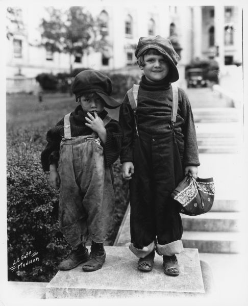 Outdoor portrait of two children wearing caps and overalls. The Wisconsin State Capitol is out of focus in the background. Caption reads: "Madison Wis. Strand theater publicity picture for motion picture 'Trouble' starring Jackie Coogan."