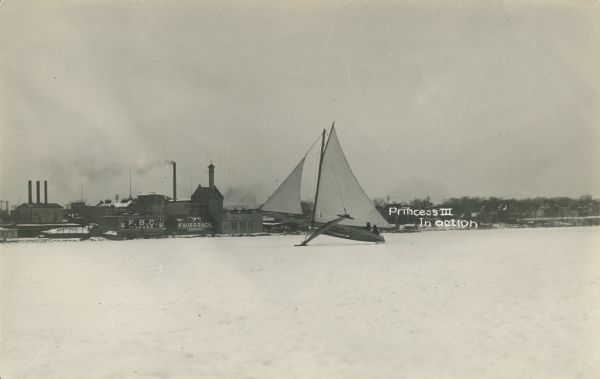 Caption on front reads: "Princess III in Action". A view across the lake towards the <i>Princess III</i> on Lake Monona with the Fauerbach Brewery in the background. Two people are on the iceboat, and it is tipped towards the right with one of the blades up in the air. The iceboat was owned by the Fauerbachs. 