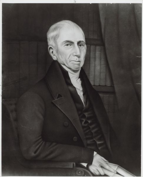 Nathan Dane is sitting in a chair holding a book with his finger between two pages. His jacket and vest are dark and his hair is white. 