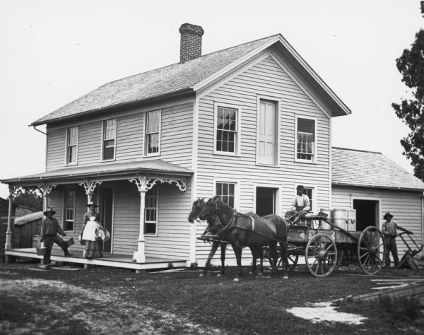 Detail from original. Original caption reads: "A farmer driving a horse-drawn wagon with milk cans. Behind him is a frame structure, possibly a cheese factory, with a porch and a full door on the second story. A man and woman are standing on the front porch of the building.  Another man is standing on the right behind the wagon.  Since this photograph was taken by Andreas Dahl, it is likely the scene was located somewhere in Dane County."