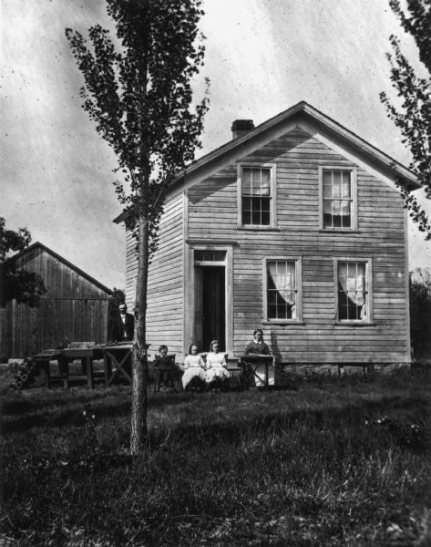 Detail from original. Original caption reads: "A couple with three children posing in a yard. The woman has a sewing machine, the man has a table saw and lathe. The two-story frame house has a glass panel above its front door, and a ladder is leaning against a tree. There is a frame outbuilding in the background. Lombardy poplars are in the foreground."