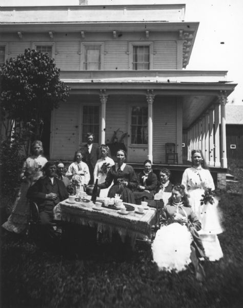Detail from original. Original caption reads: "A large family group is gathered around a table for coffee. The women are holding bunches of flowers, and behind them is a large bracket style house with a porch on at least two sides. A farm building is in the far background on the right."