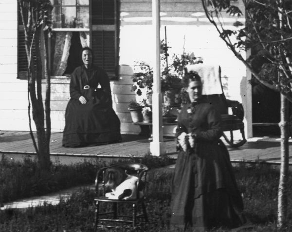 Detail from original. Original caption reads: "Five women posing in a yard. There is a man on the left standing and holding a shovel. Two children are standing on the porch roof of the frame house, which has several additions. The porch is continuous along two sides of the house."