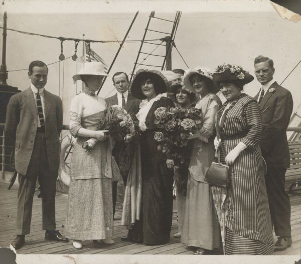Actress Pearl White stands on the deck of the RMS Olympic with four other women and four men.  All are dressed very nicely and two of the women hold large bouquets of flowers.