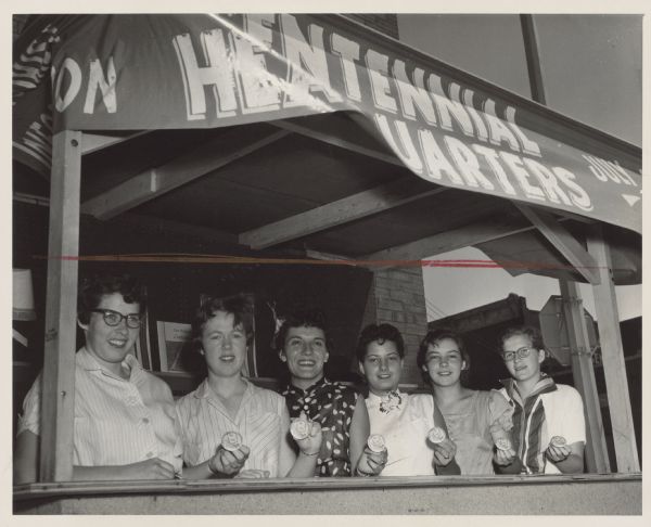 Six women posing together, all holding identical buttons. Above them is an awning banner that reads, in part: "Centennial Headquarters." Caption reads: "<b>DISPLAYING</b> Centennial buttons are six queen candidates. From left they are Mary Jo Casey, Peg Ward, Marie Sias, Arloa Kunze, Gail Pederson, and Nila Elkin. Other candidates not shown are Joan Dittman, Ruth Parnell, Janice Flatland, and Jeanie Gowan."