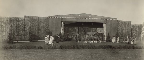 View across lawn towards people standing in front of a large theater stage and backdrop with trees drawn on it. Some are costumed as soldiers and some as Native Americans. Caption reads: "Green Bay, Wis.  July 1934 Tercentennial celebration of the discovery of Wisconsin by Jean Nicolet Historical pageant at Fort Howard".