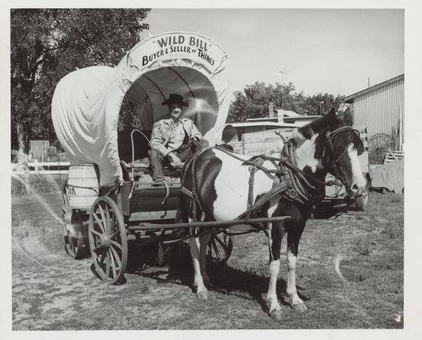 A man in a cowboy hat and fringe jacket posing on a covered wagon. The wagon has a sign that reads: "'Wild Bill' Buyer & Seller of Things, Albertville."

An attached news article reads: "ALBERTVILLE, WIS. - When the Centennial parade rolls down the main street at Colfax at 2 p.m. today, there will be, among the many units, one particularly appropriate to the occasion. That is a covered wagon from nearby Albertville.
The wagon is the handiwork of Bill Nelson of that village, and it was just completed this summer just in time to enter the Rodeo parade at Eau Claire and take first place in the 'most colorful' division and handsome trophy. Nelson said that he had to build the covered wagon 'from the bottom up,' the bottom, or chassis, being a very old light duty wagon with wooden wheels. The job wasn't easy. The builder first went to the Eau Claire library and did a little research so he would get things right. Getting things right wasn't easy either.
'I used green lumber so that it would warp and look authentic,' Nelson said. The coloring was an even greater problem. Nelson finally found that a shingle stain called bungalow brown was just the thing. One coat made the boards look old. A second coat made the boards look new and he had to scrape the first experimental swipes of that off again.
Nelson installed a feed box at the end of the wagon in the traditional manner. He said that this was used either to carry--"
[end of clipping]