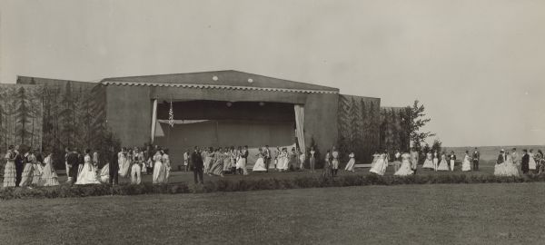 View across lawn towards people in formal attire standing in a field, in front of a tent stage. Along the tent are large panels with drawings or paintings of trees. Caption reads: "Fort Howard Ball, Green Bay Centennial, from Lillian Krueger."