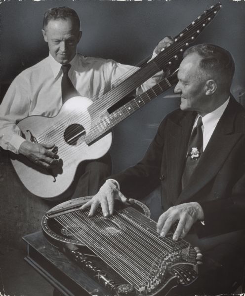 A man playing a two-necked contraguitar while another man is playing a zither. Caption reads: "<b>A bass guitar and zither</b> were played by Hans Glieber (left), 2121 W. Raleigh av., Glendale, and Joseph Greiler, 3147 N. 39th st., at the winter festival of the United Bavarian Societies, Inc., Sunday at Jefferson hall, 2617 W. Fond du Lac av."