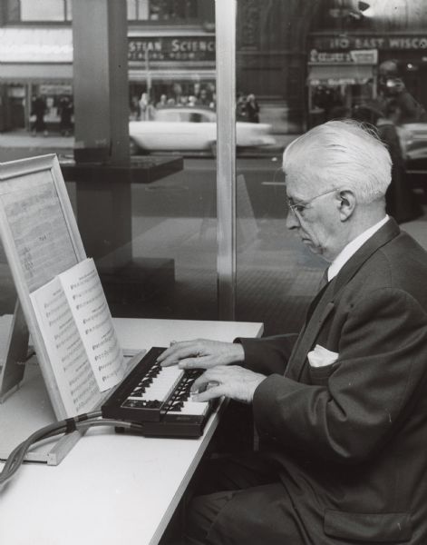 A man is playing a small double keyboard, from which tubes are extending. Behind him are large windows looking out towards a street scene. Across the street is a storefront with is a sign for a Christian Science reading room, and another has a sign for 110 East Wisconsin Avenue. Caption reads: "Anton Brees at the carillon keys."