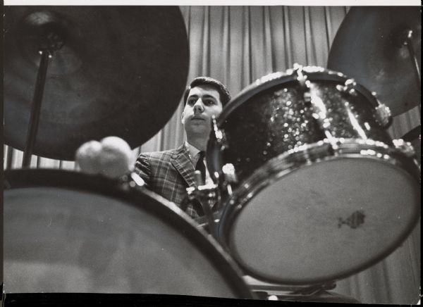 View of a young man, surrounded by drums in the foreground. Caption reads: "<b>A Whitefish Bay youth,</b> Robert Bealmear, of 5324 N. Kent av., beat the drums as Groove, Inc., a 17 piece jazz stage band, played at a meeting of the Milwaukee Jazz society at Radio City."