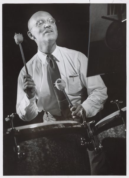 A man is using two mallets on a drum while looking over at a music stand. Caption reads: "<b>A timpani player rehearsed</b> with the Jewish Community Center orchestra Wednesday night at the center, 1400 N. Prospect av. He is Dr. Louis H. Kretchmar of 2821 E. Menlo blvd., Shorewood. It was the orchestra's first rehearsal of the season."