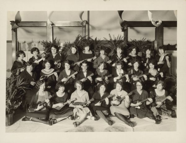 Group portrait of 24 women and one man sitting and holding ukuleles. Caption reads: "Young women's ukulele orchestra at French Battery and Carbon Co. party held at East Side Club, Madison, Wis., Nov. 30, 1925."