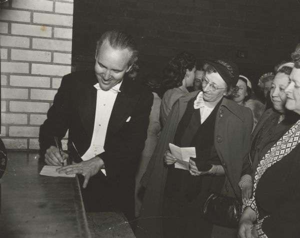 A man is signing a piece of paper, while three women are looking on. Caption reads: "Prof Gunnar Johansen during benefit concert for swimming pool campaign at Mt. Horeb."