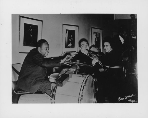 Three men and one woman playing instruments in a band. Framed photographs are hanging behind them. Caption reads: "Jimmy [sic] Noone (Negro) orchestra, ca. 1941. L. to r.: Warren 'Baby' Dodds, drums; Jimmy Noone, clarinet; Maida Roy, pianist. Man at bass viol. not identified. Photo taken at Chicago, ca. 1941. Copied from a photo loaned by Warren Dodds, Chicago (via Larry Gara, Madison.) See Dodds 'tapes' in Manuscripts Room."