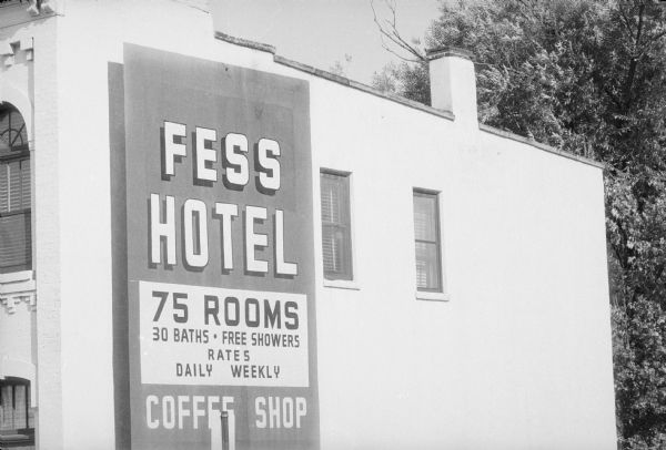 View towards the front right corner of the Fess Hotel, and the sign painted on the right side of the Fess Hotel that reads: "Fess Hotel, 75 Rooms 30 Baths • Free Showers • Rates Daily Weekly, Coffee Shop". 