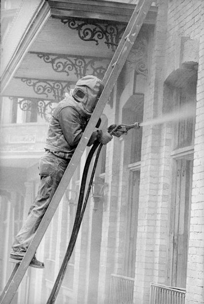 View of a man standing on a ladder and sandblasting the brick facade of the Fess Hotel. He is wearing a protective hood, and is working on the right side of the building near windows under a balcony.