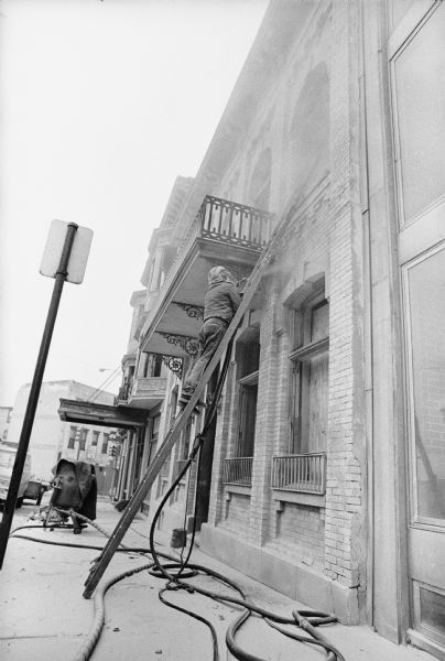 View from sidewalk of a man standing on a ladder and sandblasting the facade on the right side of the Fess Hotel.