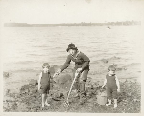 Actress Pearl White stands on the beach with two small boys — Billie and Buddie — one of whom is her nephew. The three are digging for clams. White wears a hat and long wading boots and holds a clam fork; one of the boys stands next to a metal pail.